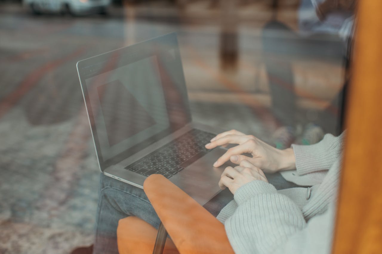 A person types on a laptop indoors, reflected through a window, wearing a comfy sweater.