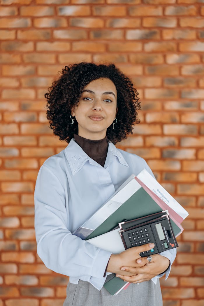 A confident woman in white long sleeves holding folders and a calculator against a brick wall.
